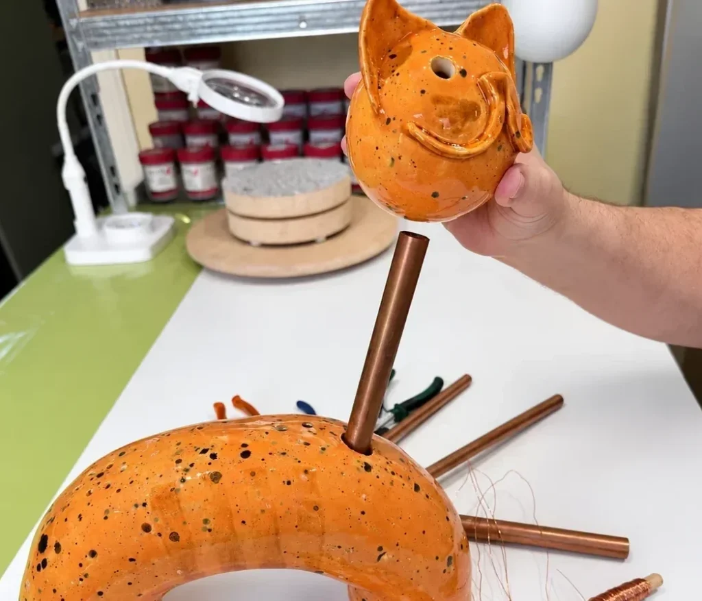 Der Kopf der orange glasierten KERAMIKATUR wird probeweise auf den noch nicht befestigten Metallhals gesetzt. Position von Hals und Kopf werden für die spätere Drehbarkeit genau abgestimmt.